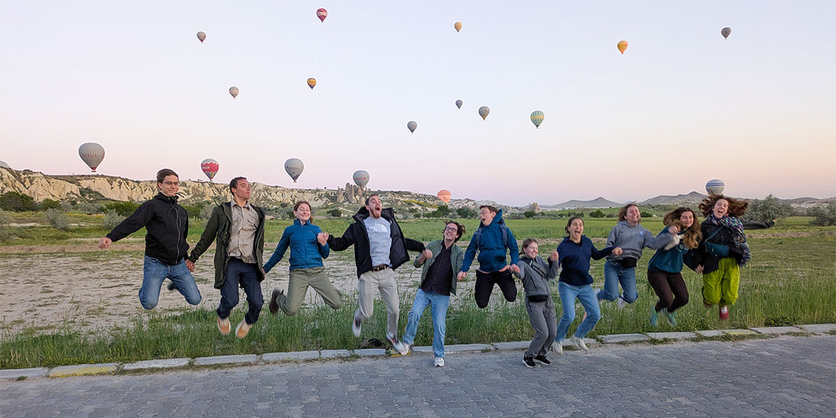 group of students jumping in the air in front of a sky full of hot air balloons