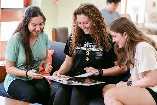 Three students sitting on a couch, smiling and looking at a book together.