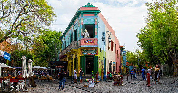 olorful street scene in 布宜诺斯艾利斯, 阿根廷, featuring a vibrant building with a statue on the balcony, cobblestone streets, outdoor cafés, and people walking.