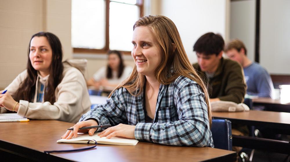 Smiling student in a plaid shirt taking notes in a classroom with other students in the background.