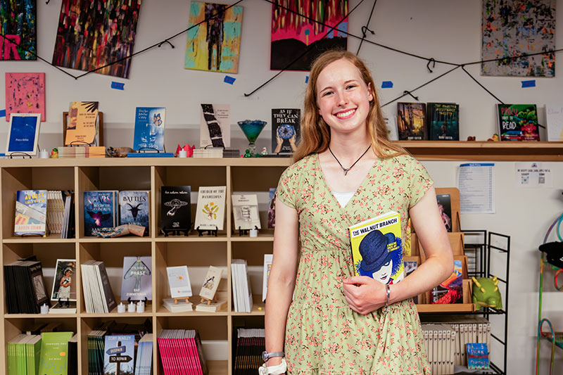 Smiling young woman in a floral dress holding a book while standing in front of colorful shelves and artwork in a bookstore or library.