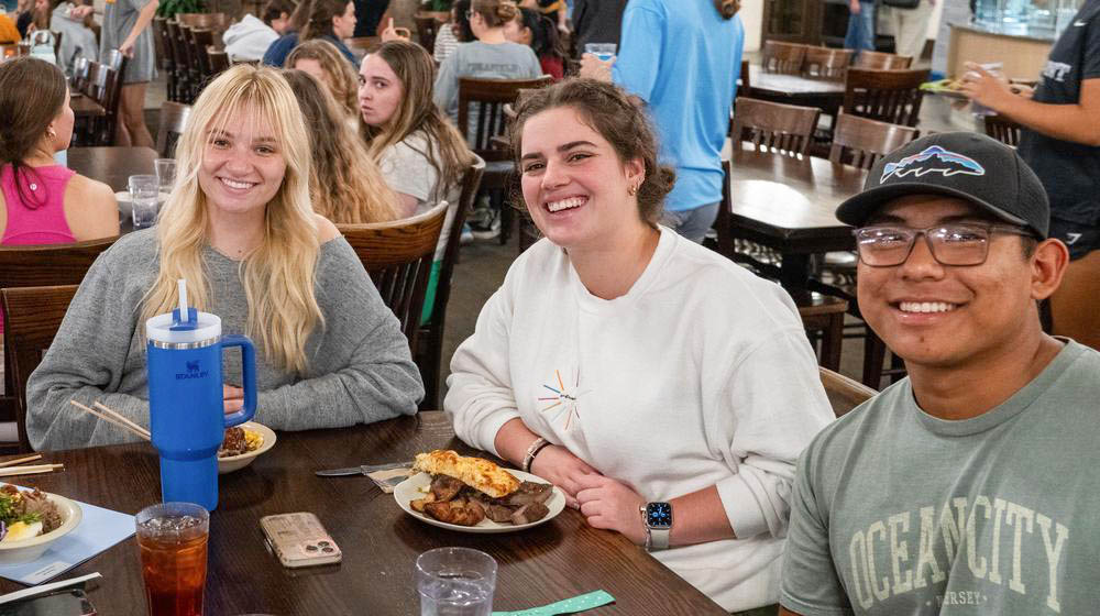 Three students smiling at a table with plates of food in a busy dining hall