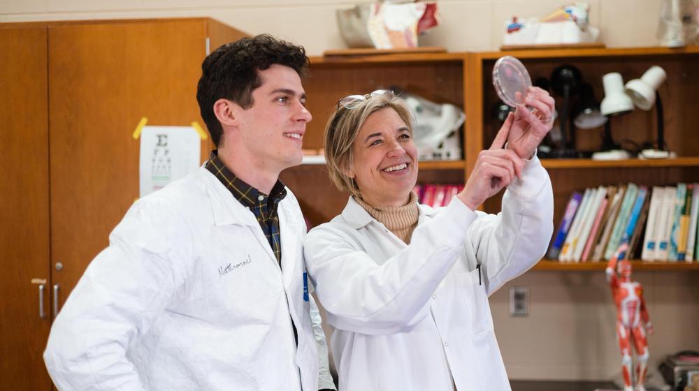 Student and professor in lab coats examining a petri dish in a science classroom.