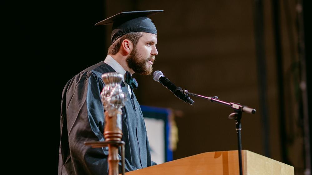 Graduate in cap and gown speaking at a podium during a commencement ceremony.
