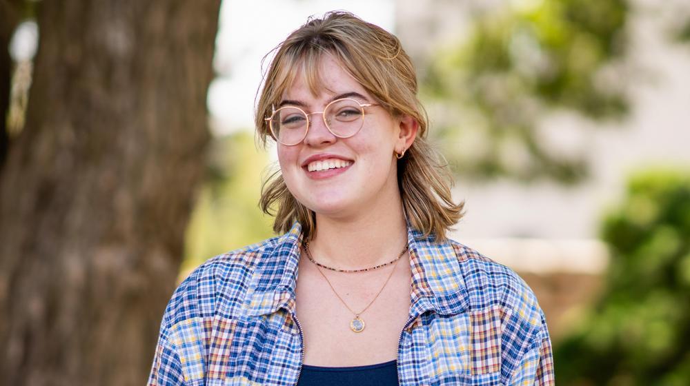 Smiling student wearing glasses and a plaid shirt poses outdoors in a sunny, tree-filled setting.
