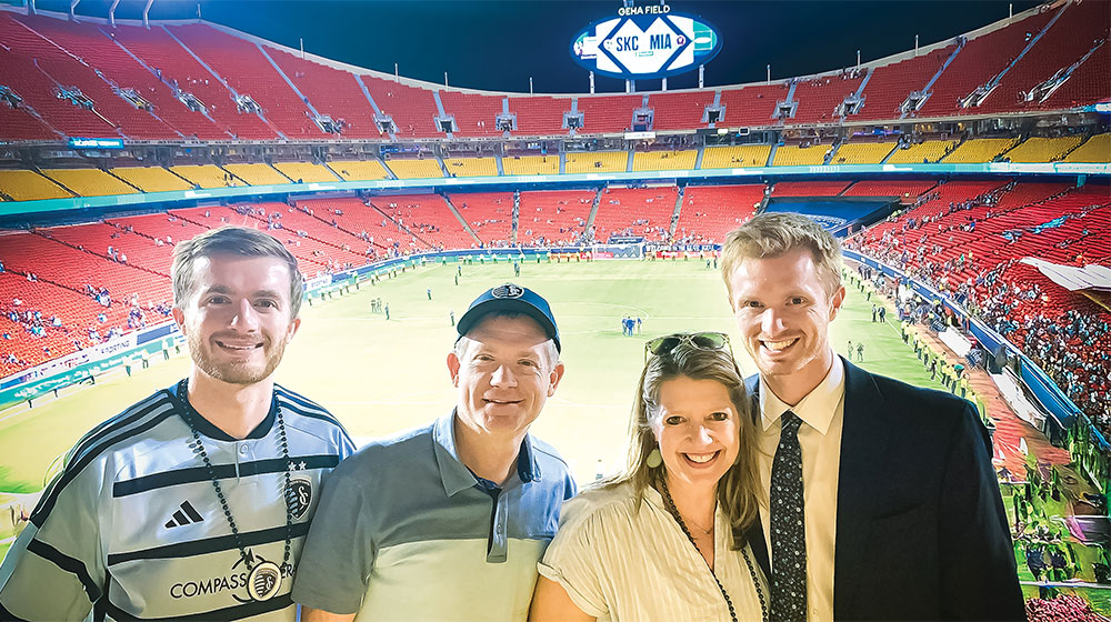 Four people posing and smiling in front of a large stadium during a night match.