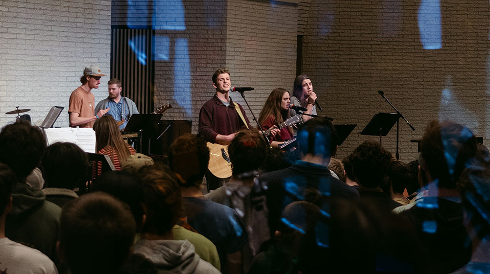 Band leading worship with guitars and vocals before an audience in a chapel-like setting