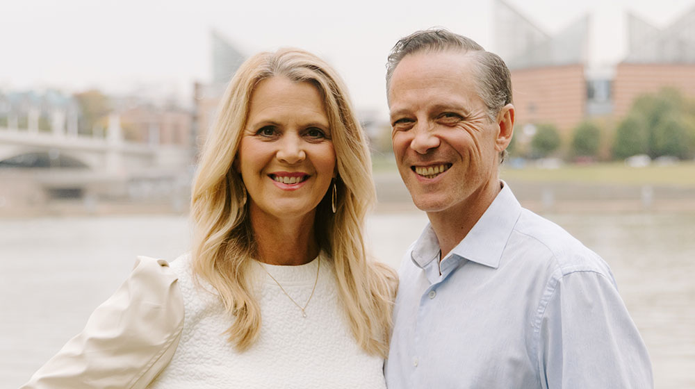 Smiling couple posing by a riverside with modern buildings and a bridge in the background.