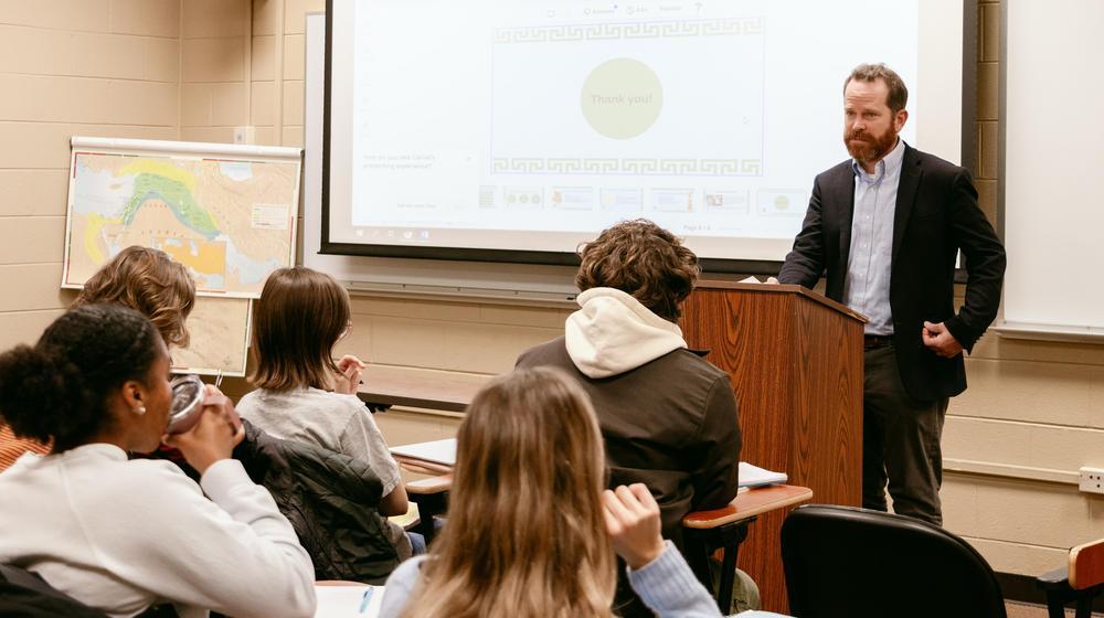 Professor standing at a podium giving a presentation to students seated in a classroom with a projected slide and map in the background.
