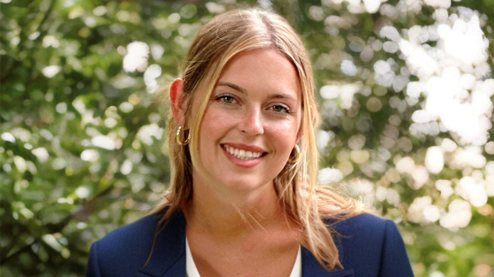 Smiling woman in a blue blazer posing outdoors with greenery in the background.