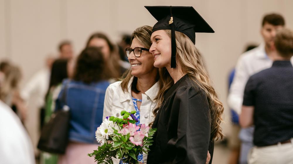 Graduate in cap and gown smiling and posing with a woman holding a bouquet of flowers at a ceremony