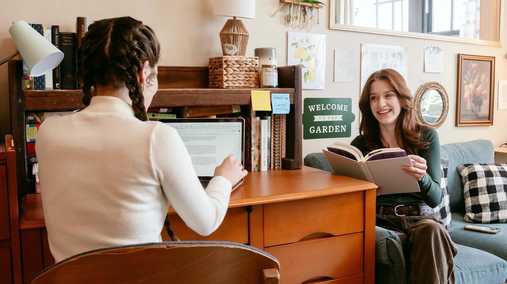 Two students in a dorm room, one working on a laptop and the other reading a book, chatting together.