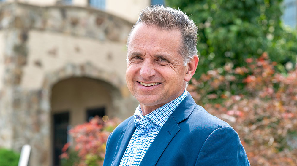Smiling man in a blue blazer standing outside in front of a stone building and greenery.