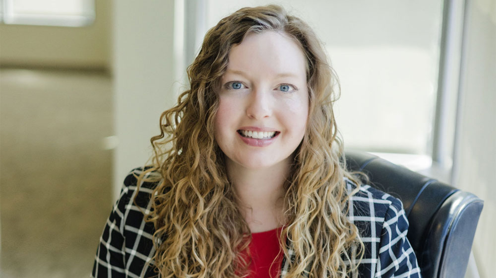 Smiling woman with curly hair, wearing a red top and a black-and-white checkered blazer, sitting in a well-lit office setting.