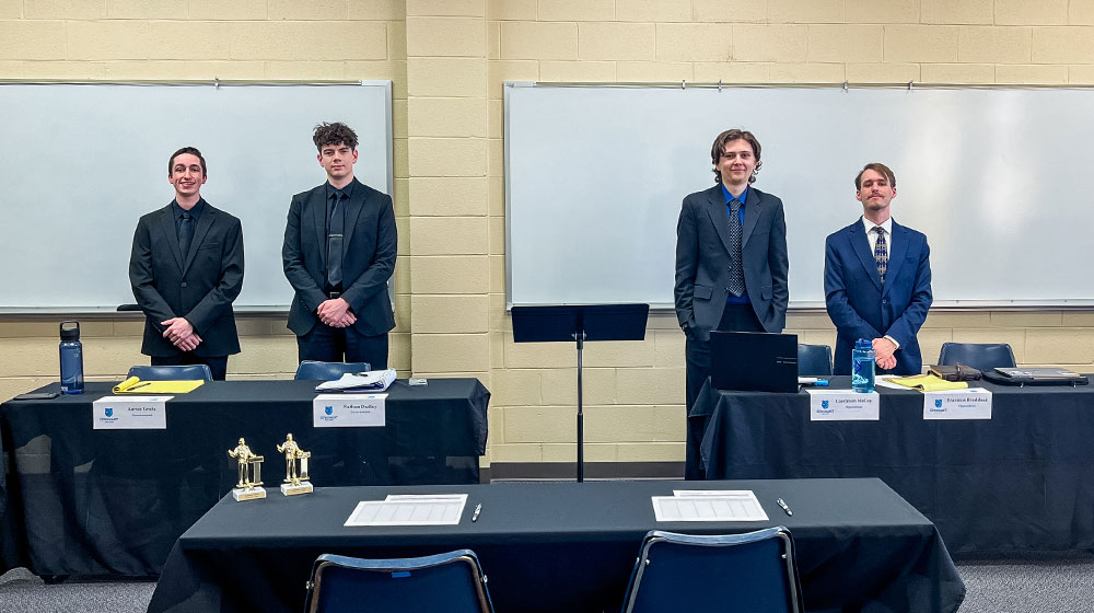 Four students in suits standing behind debate tables with trophies in front.