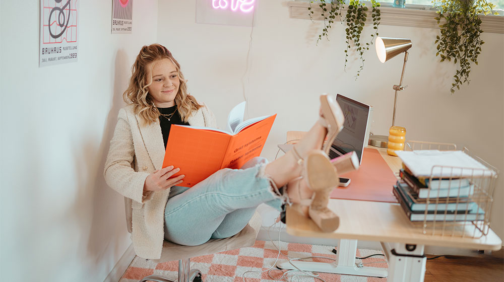 Woman seated at a desk in a home office, legs propped on the tabletop, reading an orange-covered book.