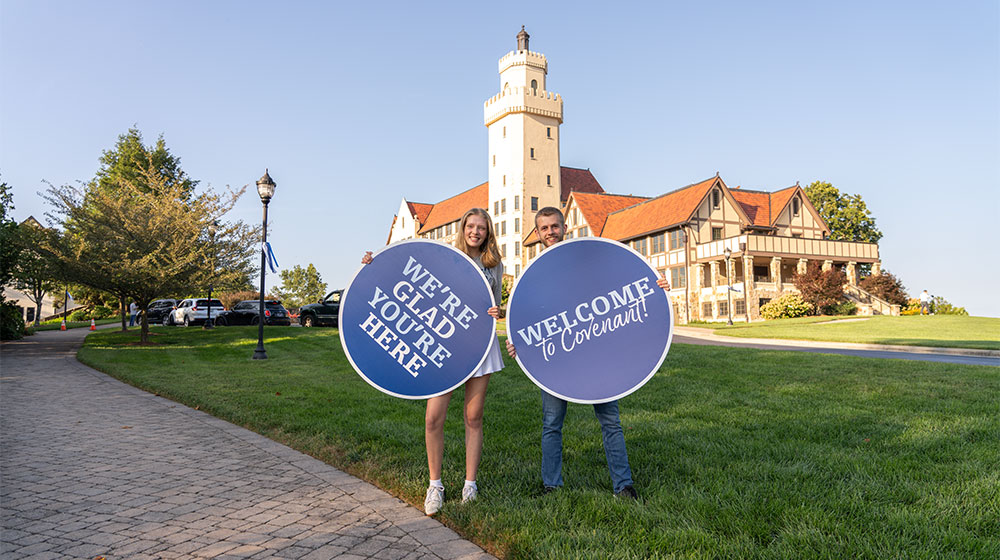 People holding circular signs reading We’re glad you’re here and Welcome to Covenant on a campus lawn in front of a castle-like building.