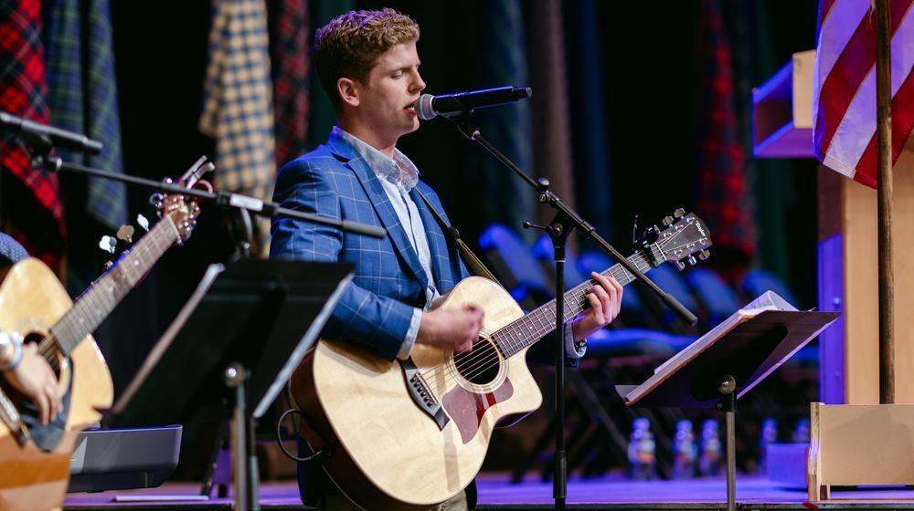 Musician in a blue blazer singing and playing guitar on stage during a ceremony, with tartan banners and an American flag in the background.