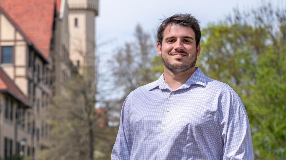 Man in a light checkered shirt smiling outdoors with a historic building and greenery in the background.