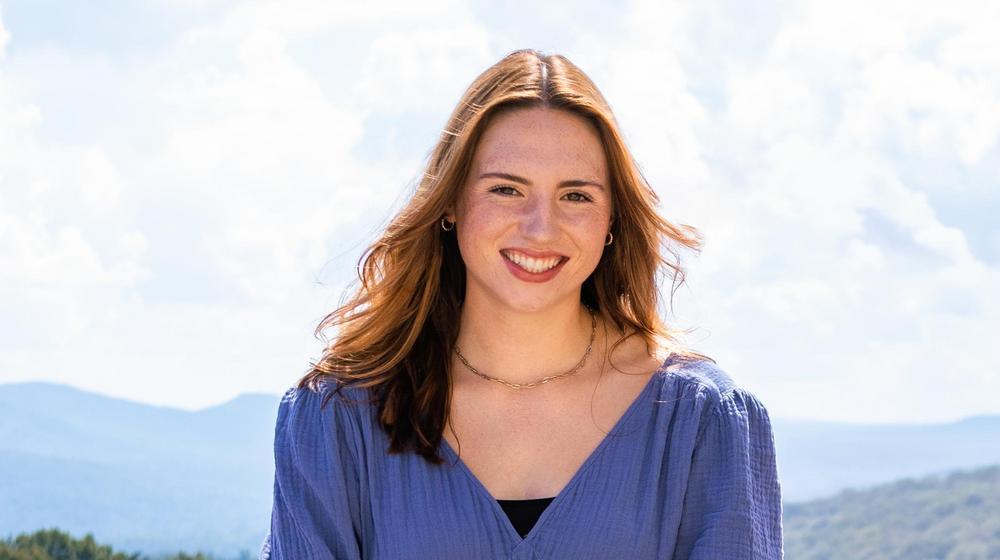 Smiling young woman with auburn hair wearing a blue blouse, with mountains and sky in the background.