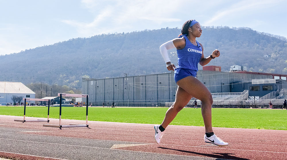 Track athlete in a blue Covenant uniform running on an outdoor track with mountains in the background.