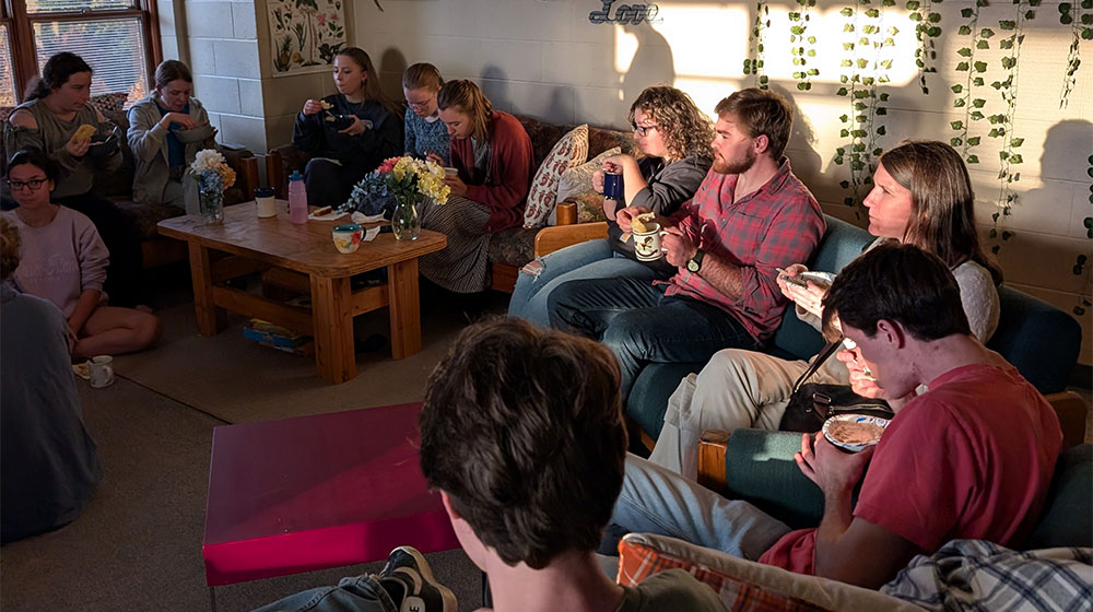 A cozy gathering of people in a warmly lit living space, enjoying food and conversation, surrounded by string lights and decorative plants.