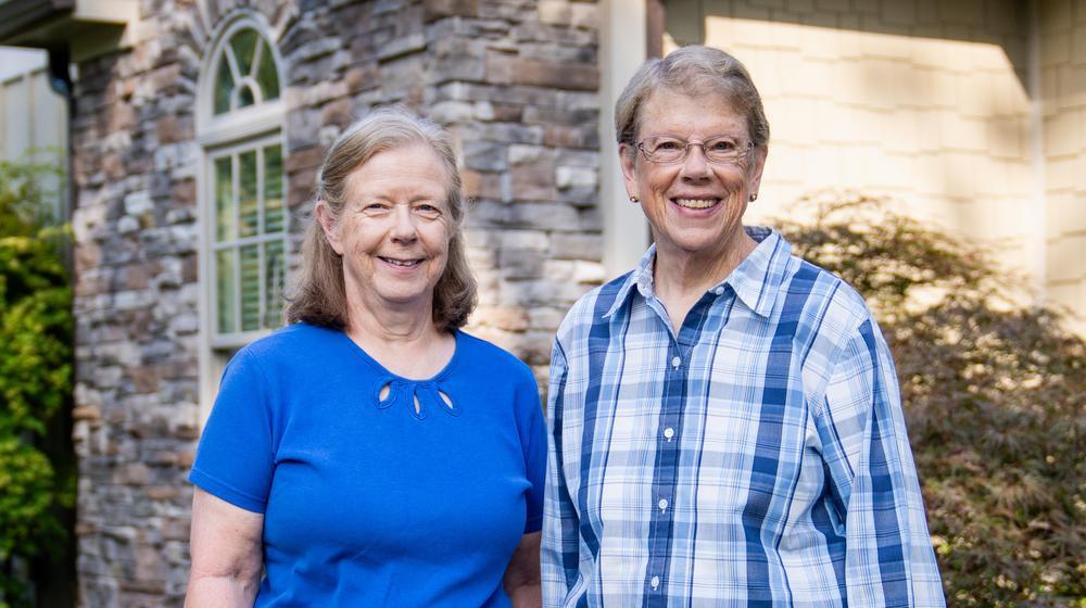 Two older women smiling outside a stone house.