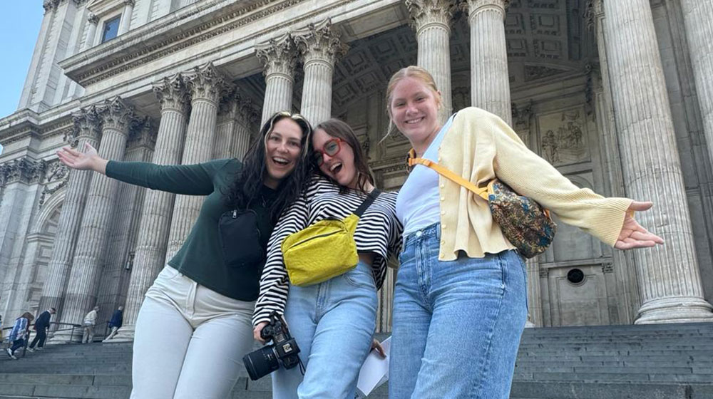 Three young women smiling and posing in front of a grand historic building with tall columns, likely St. Paul's Cathedral in London. 的y are dressed casually, wearing crossbody bags, and one is holding a camera, capturing a moment of their travels.