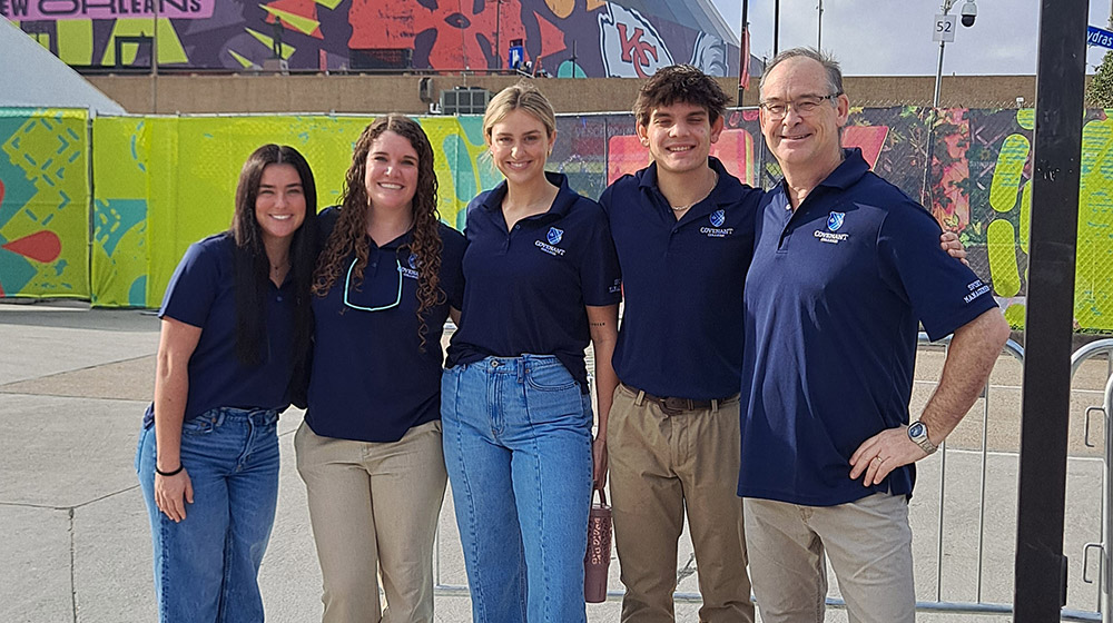 Group of five people in matching navy Covenant-branded shirts posing together outdoors in front of colorful fencing