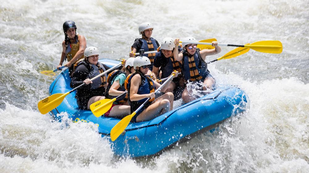Group of people wearing helmets and life jackets whitewater rafting in a blue inflatable boat.