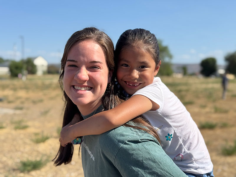 Smiling young woman giving a piggyback ride to a cheerful little girl outdoors on a sunny day.