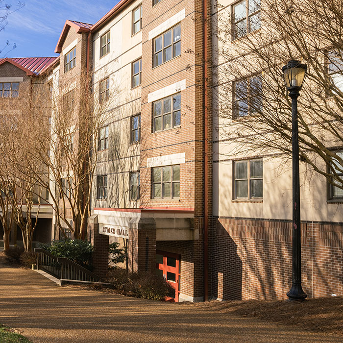 Exterior view of Rymer Hall, a multi-story dorm building with a brick facade and windows, framed by tall trees and a lamppost.
