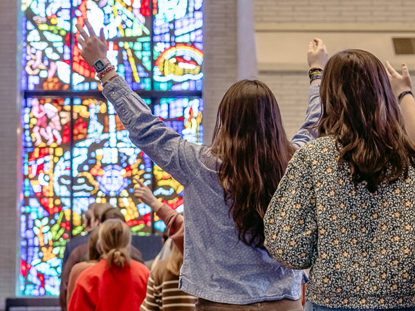 Two individuals raising their hands in worship in front of a vibrant stained glass window.